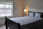 Cozy bedroom with a double bed, lamp, and window view at Stone Farm Apartments in Lebanon, NH.