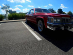 1997 Cadillac DeVille in candy apple red parked beside a lake, showcasing chrome wheels and a sleek design.