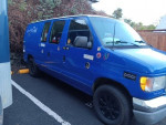2001 Ford Econoline 150 hippie camper van with blue body and stickers, parked in a driveway.