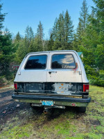 Rear view of a 1989 Chevy Suburban with peeling paint, parked in a forested area, showcasing its project potential.