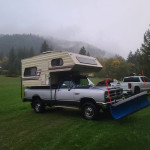 1989 Dodge Ram W250 4x4 with camper and snow plow parked on grassy field in misty mountains.